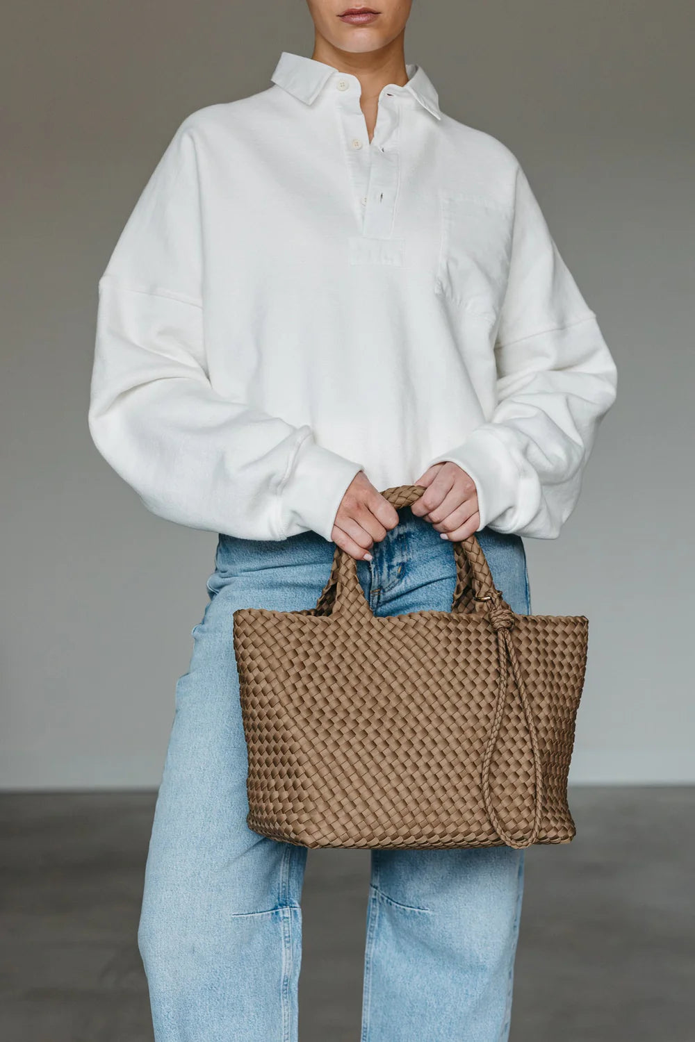 Person holding a woven brown bag wearing a white shirt and blue jeans against a neutral background
