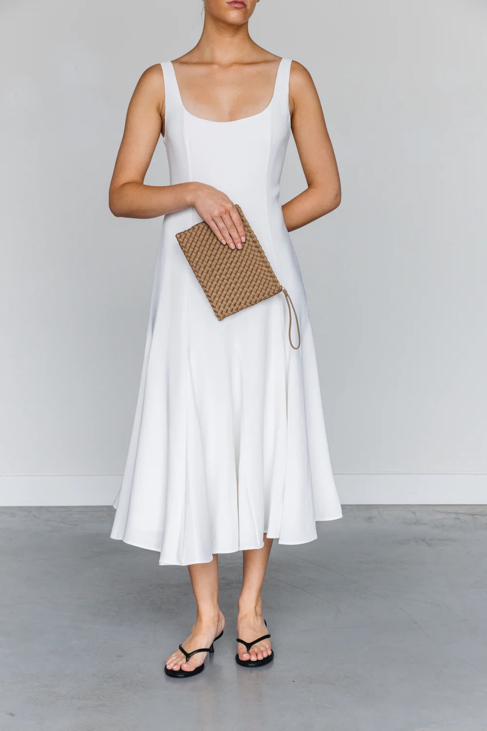A woman wearing a sleeveless white midi dress, holding a brown woven clutch, and wearing black flip-flop sandals against a light gray studio background.