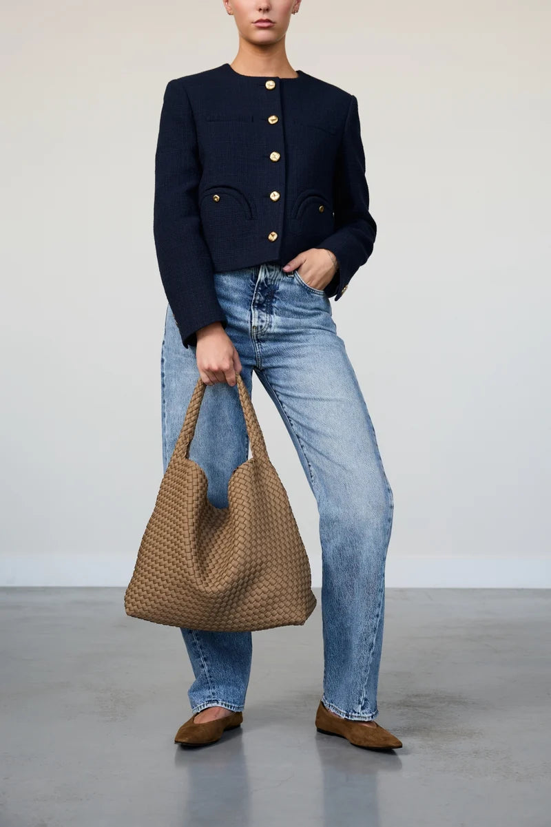 Person wearing a navy jacket and blue jeans, holding a woven brown bag against a plain background