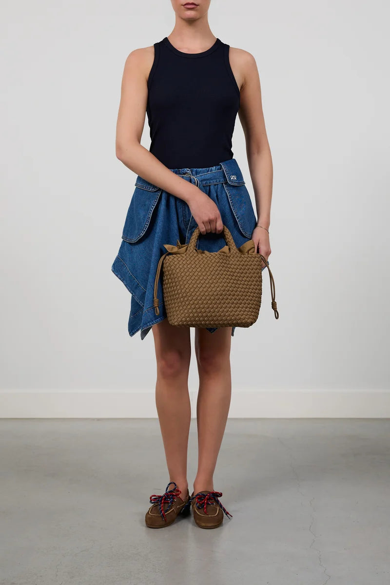 A model wearing a black sleeveless top, a blue denim skirt, and brown lace-up shoes holds a brown woven basket-style handbag against a plain light gray studio background.