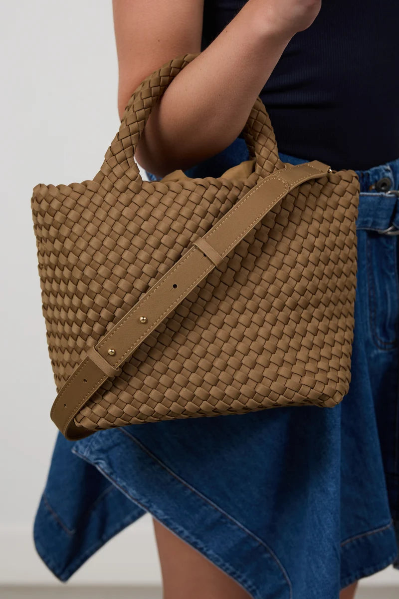 A woven brown tote bag with a matching shoulder strap, carried by a person wearing a dark top and blue denim skirt against a plain light background.