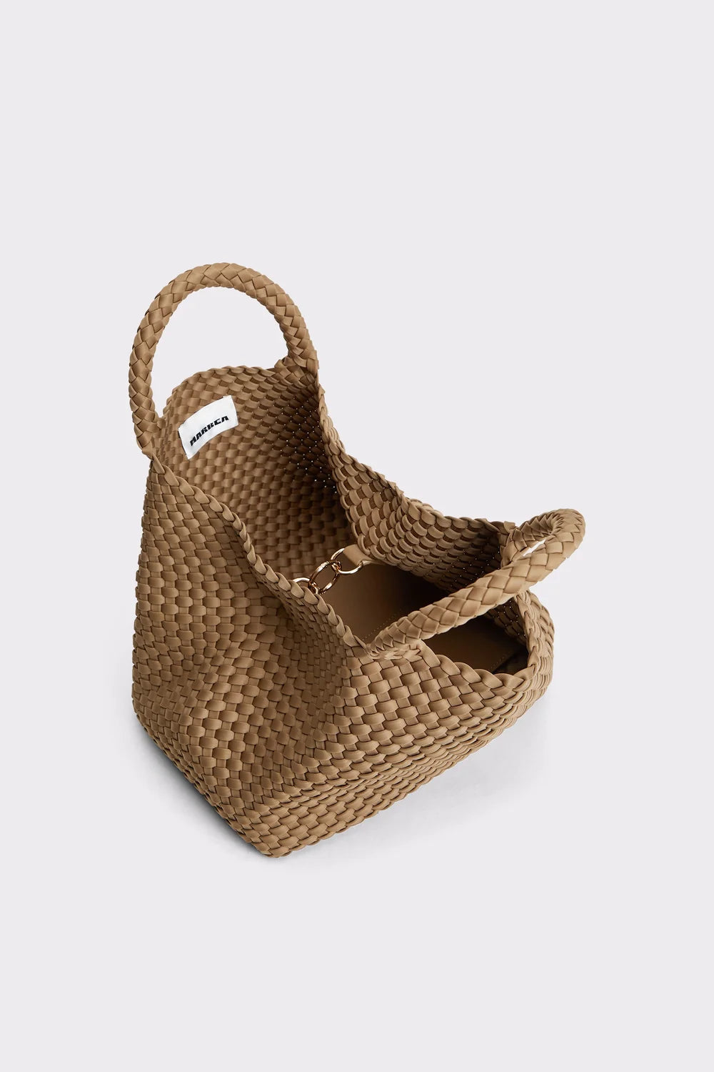 A woven straw tote bag in a natural tan color, featuring two top handles and a small white brand label on the side, photographed on a light gray background.