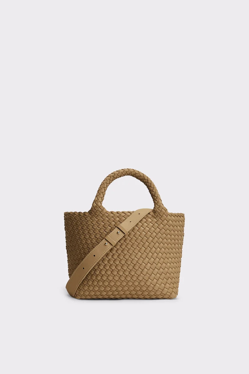 A woven straw tote bag in a natural tan color, featuring two top handles and a matching woven shoulder strap with a silver-tone buckle, set against a solid light gray background.