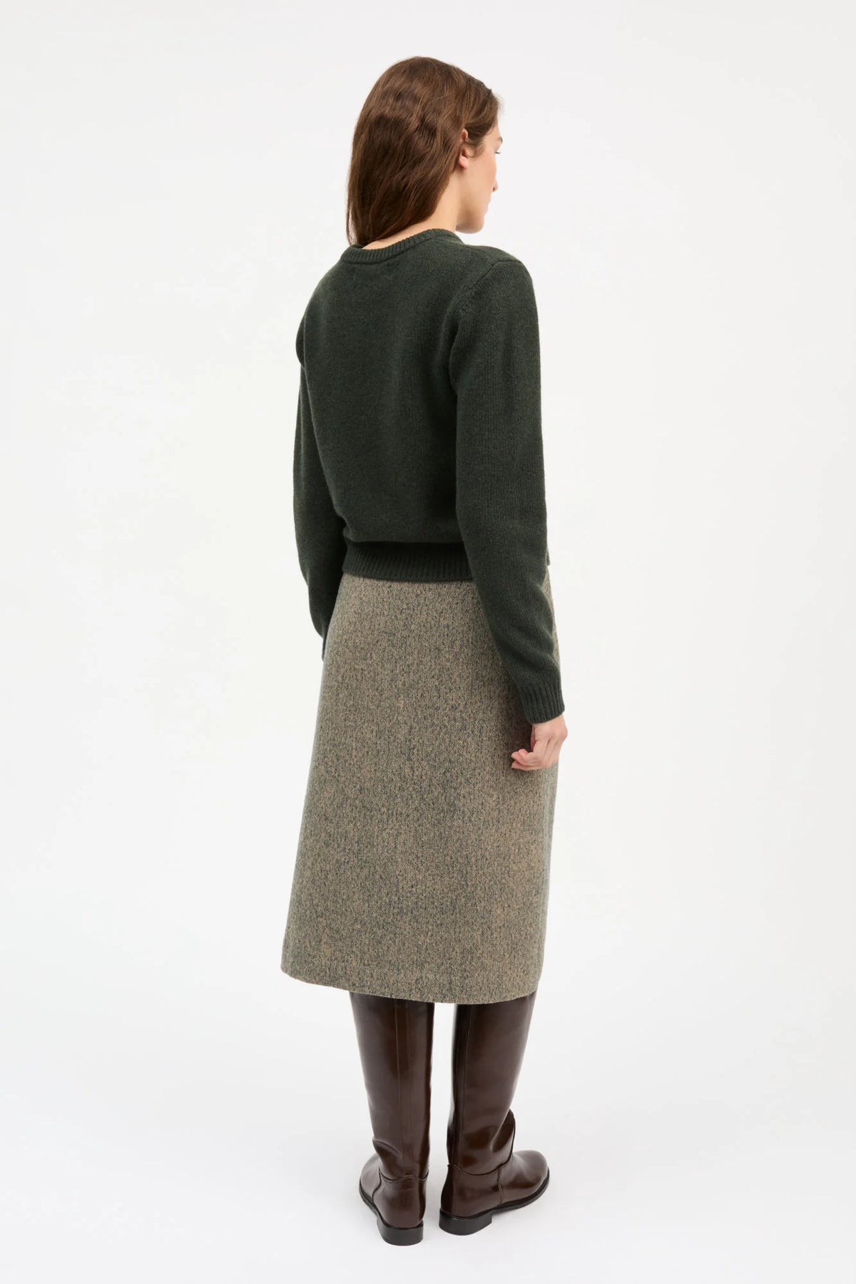 A woman shown from the back wearing a dark green long-sleeve knit sweater, a brown and beige herringbone tweed midi skirt, and dark brown knee-high boots, against a plain white studio background.