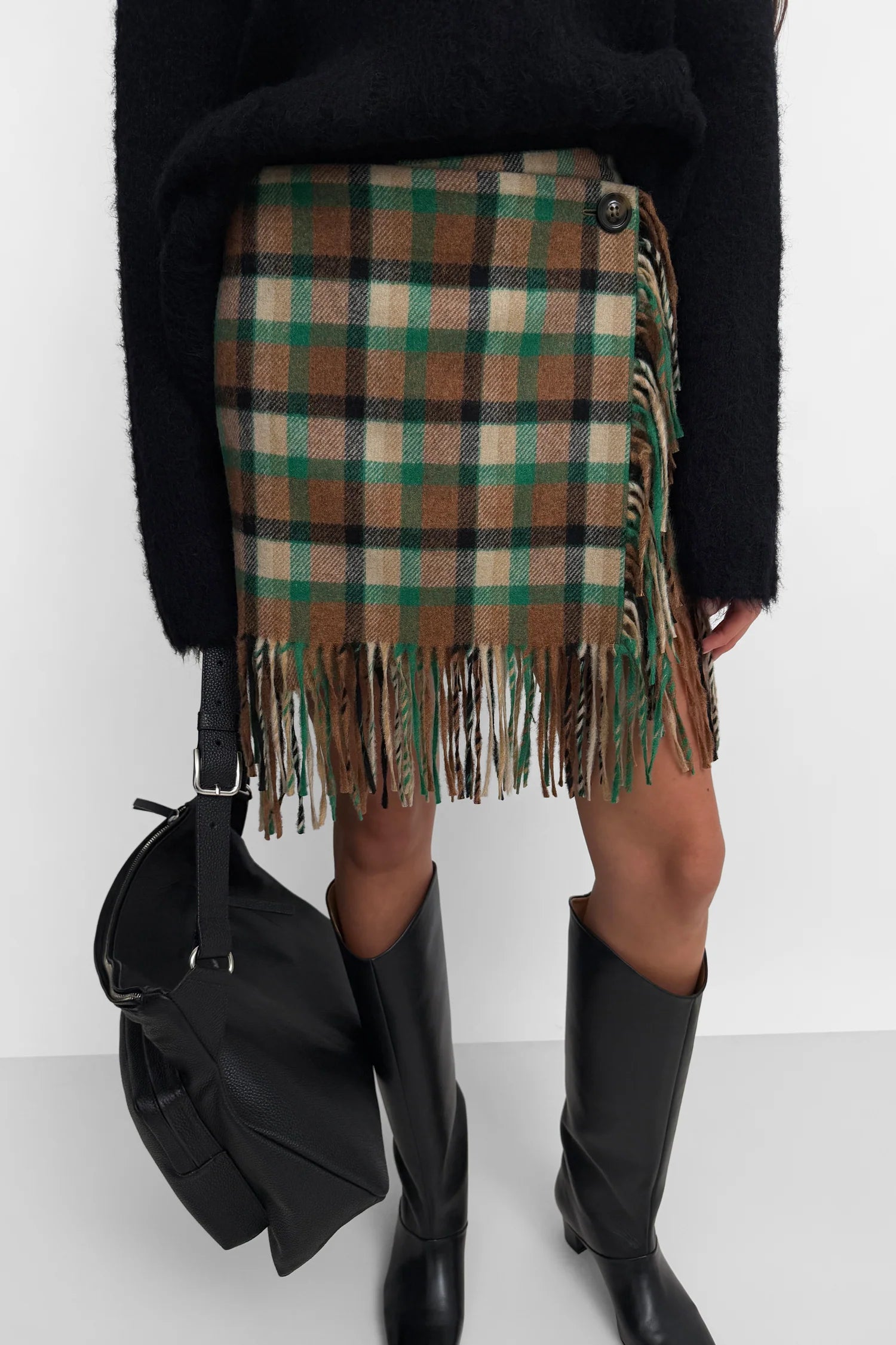 A model wearing a brown, green, and beige plaid wrap skirt with fringe trim, styled with a black fuzzy sweater, black leather knee-high boots, and a black shoulder bag against a white studio background.