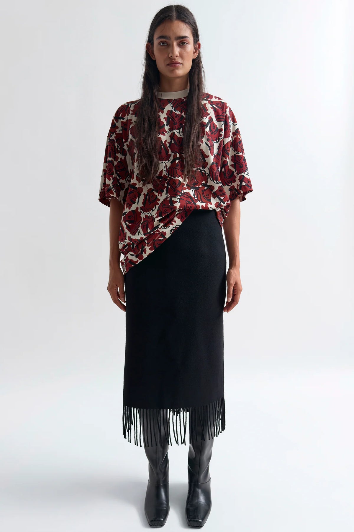 A model stands against a light gray studio background wearing a short-sleeve asymmetrical top with a red, black, and beige floral print, paired with a black midi skirt featuring fringe at the hem and black knee-high boots.