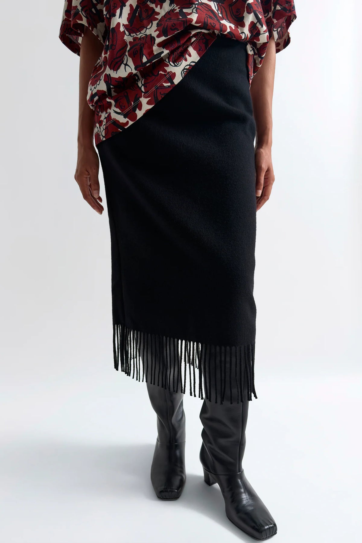 A model is shown from the waist down wearing a black knit fringe skirt, black leather knee-high boots, and a red and black floral blouse, set against a plain white studio background.