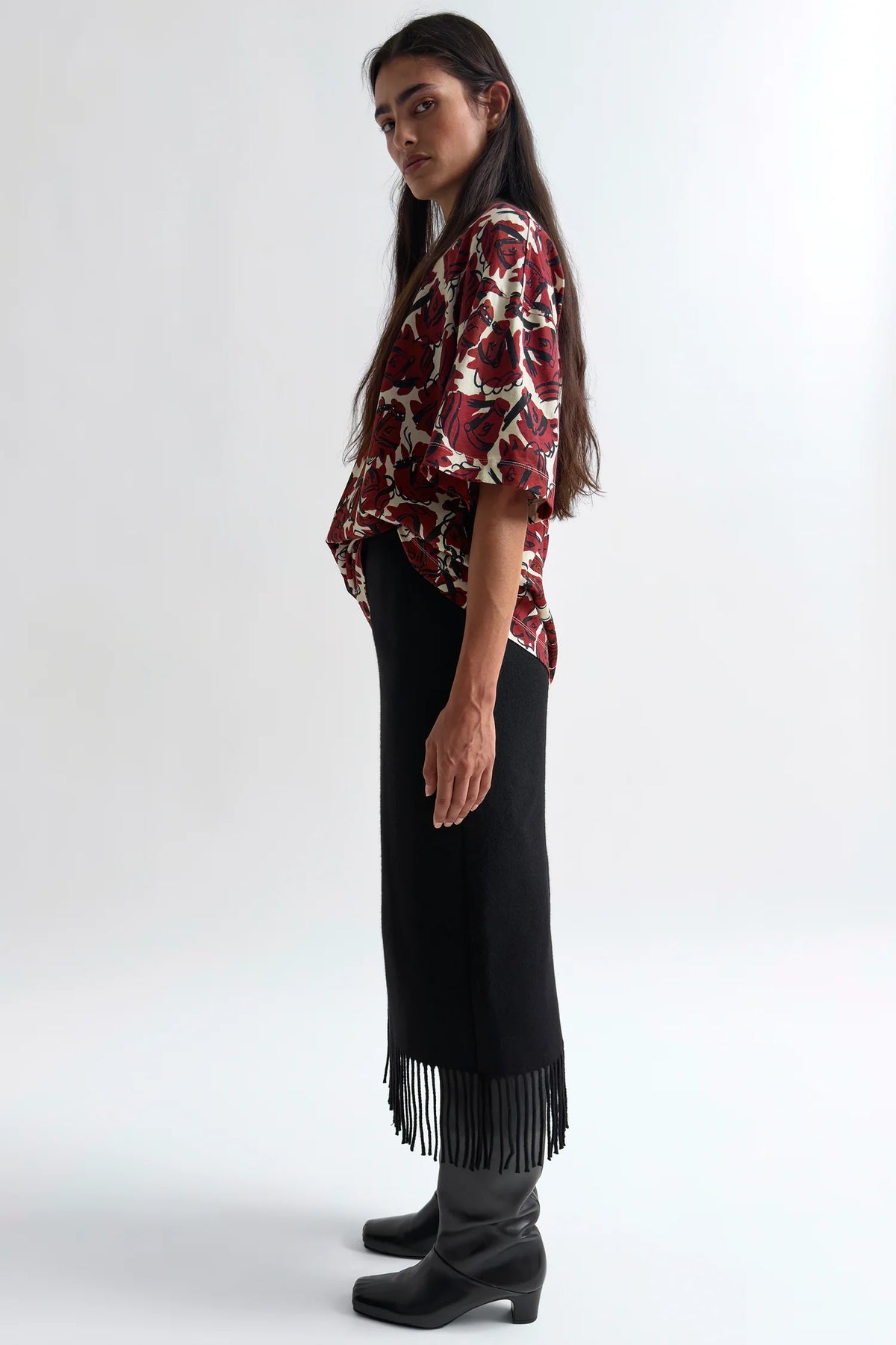 A woman stands in profile wearing a red, black, and white patterned short-sleeve blouse, a long black skirt with fringe at the hem, and black heeled boots against a plain light gray studio background.