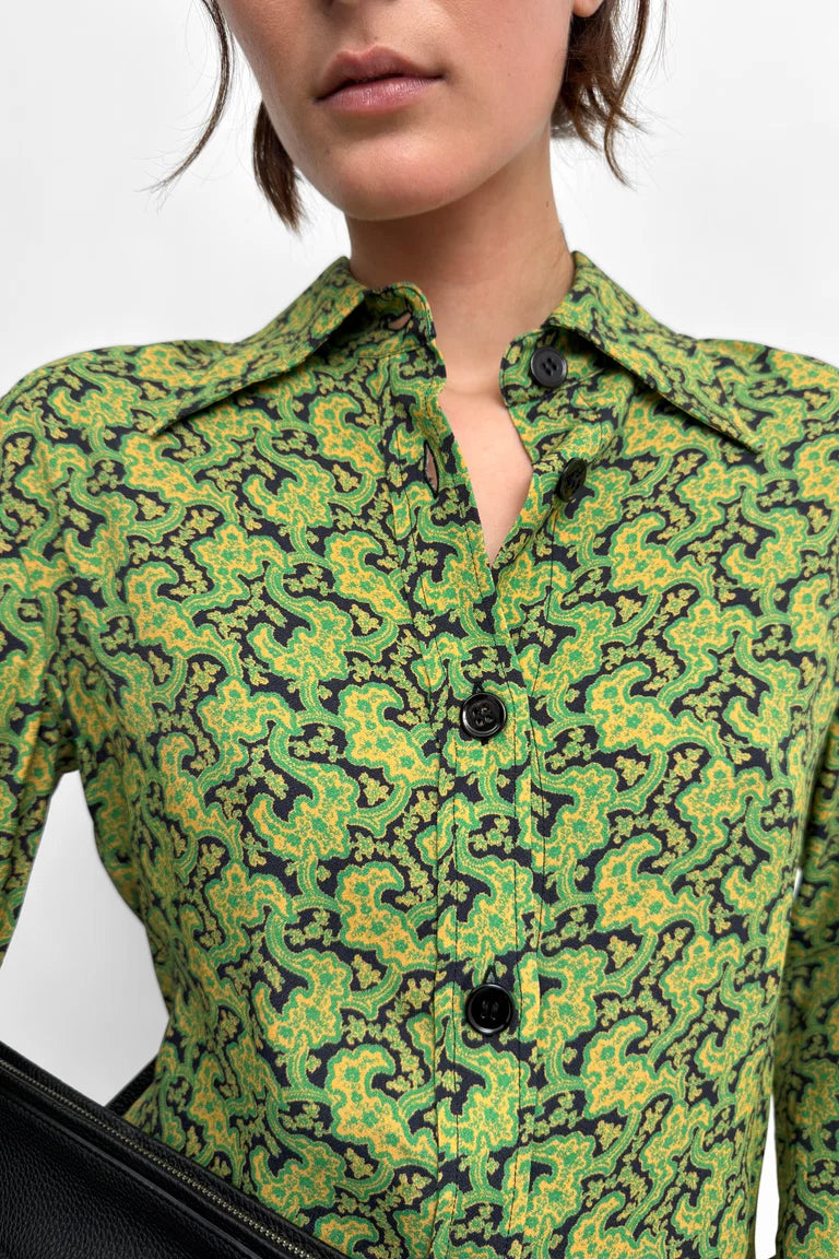 A close-up of a model wearing a green and black patterned button-up shirt, with a black leather bag partially visible in the lower left corner against a light gray background.