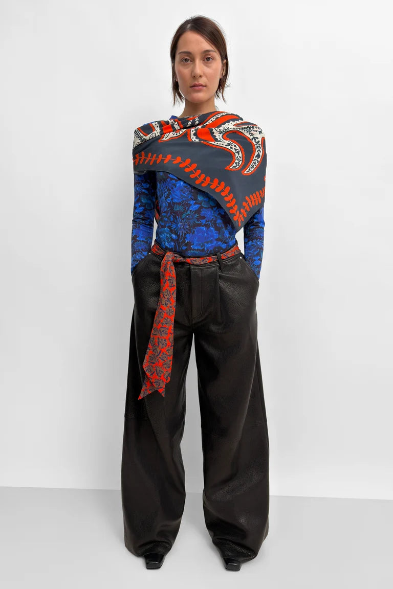 A model stands against a light gray studio background wearing a long-sleeve top with a bold blue and black paisley pattern and a red-and-black draped shawl across the shoulders. The outfit is completed with black wide-leg pants and a matching red-and-black patterned belt tied at the waist.