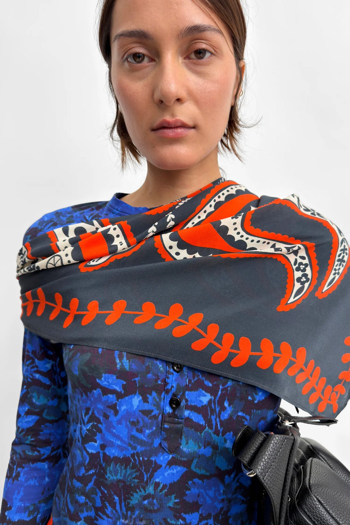 A close-up of a model wearing a dark gray scarf with a bold orange and white graphic pattern, styled over a blue floral long-sleeve top.