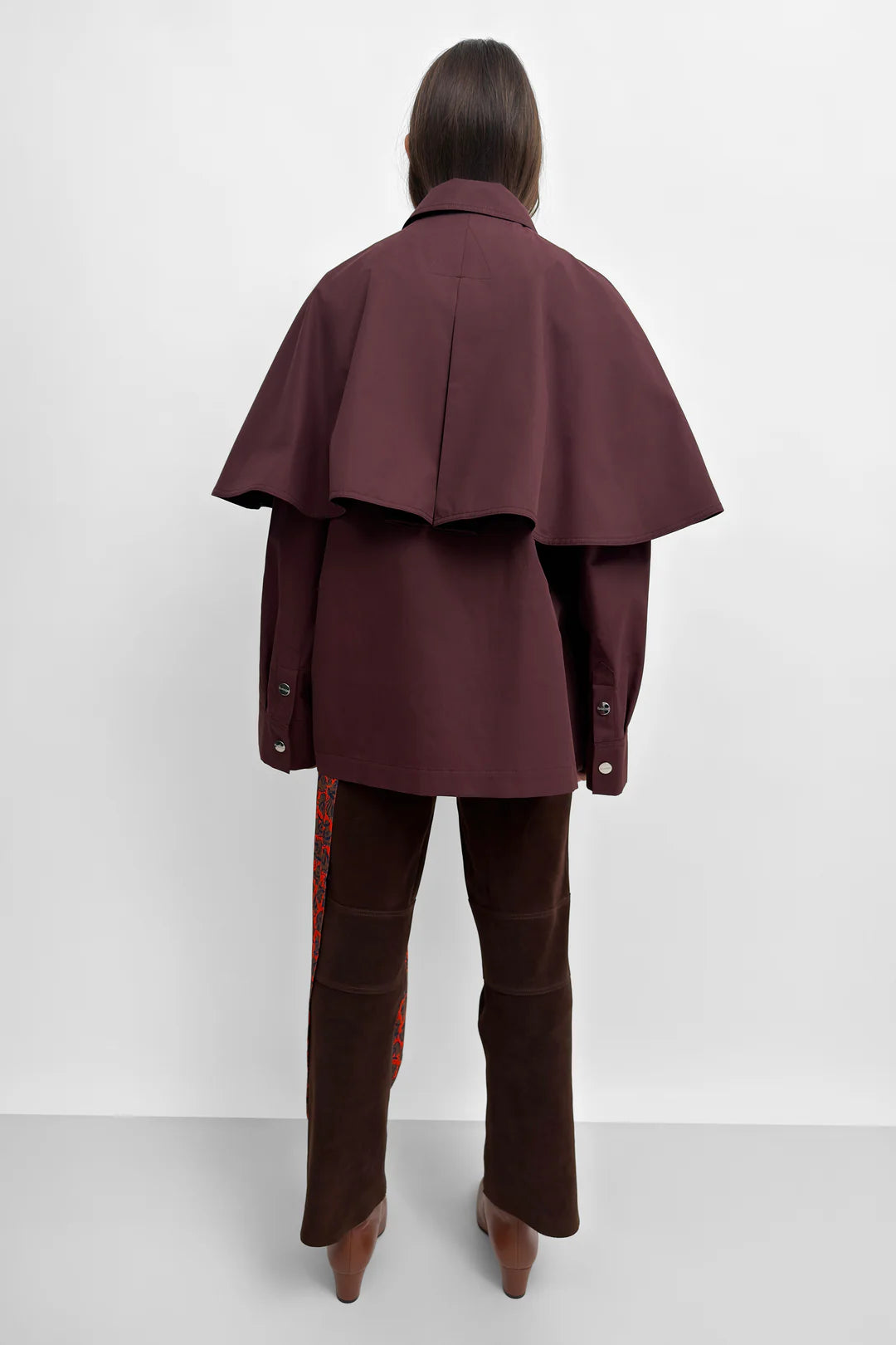 A model shown from the back wearing a dark burgundy cape jacket and matching dark brown trousers with patterned side panels, set against a plain light gray studio background.
