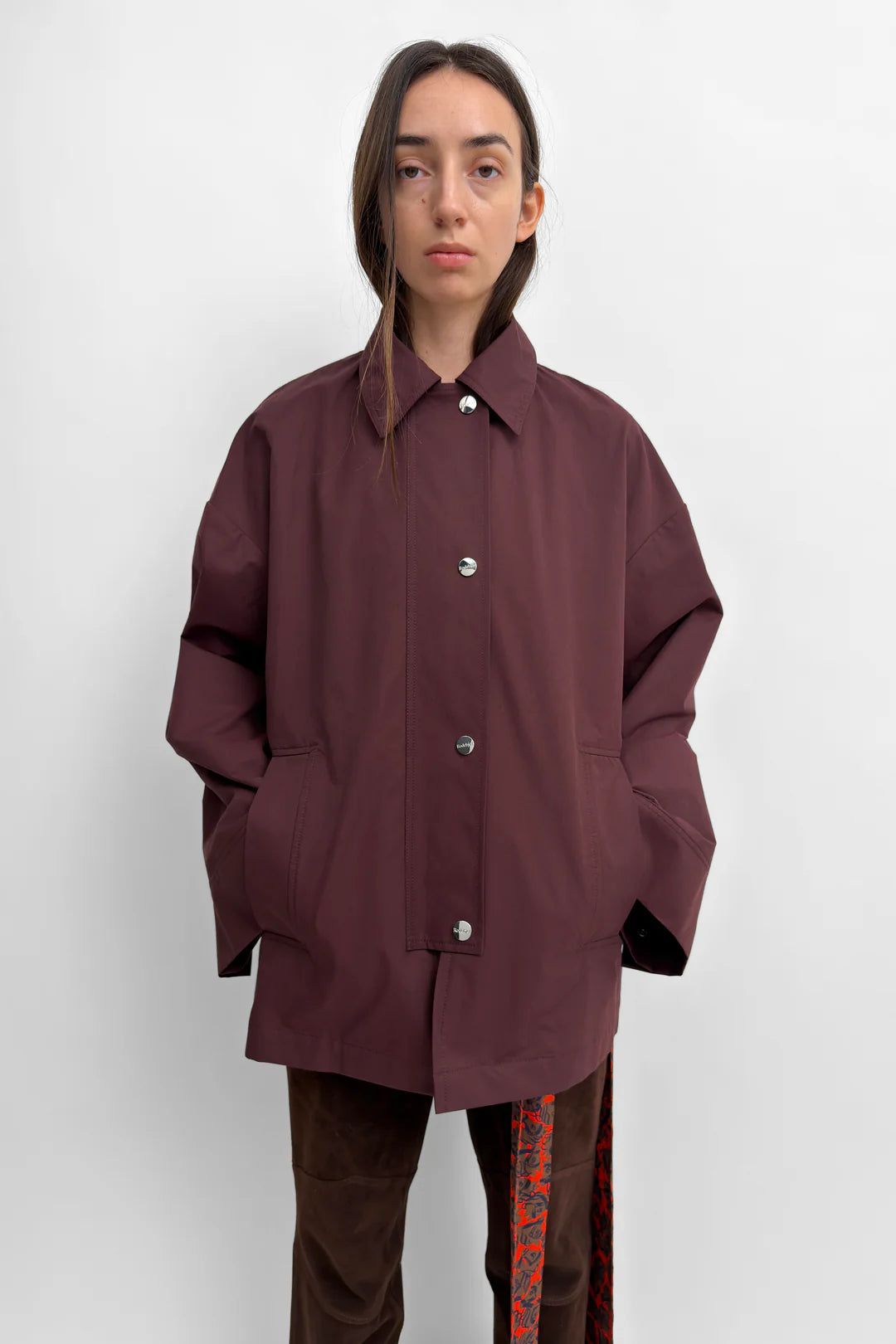 A model wearing an oversized, dark plum-colored button-up shirt jacket with silver buttons, paired with brown pants, against a plain light gray background.