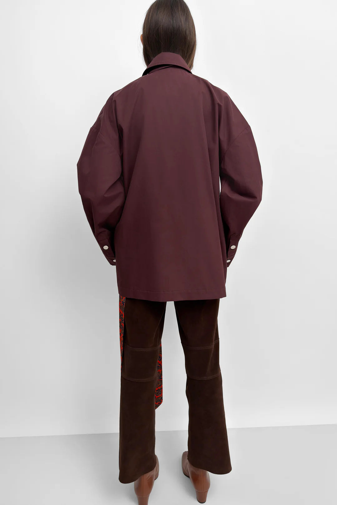 A model stands against a light gray studio background wearing a dark burgundy cape-style jacket with wide draped sleeves, dark brown trousers, and brown boots. A patterned red and green fabric hangs from the side of the pants.