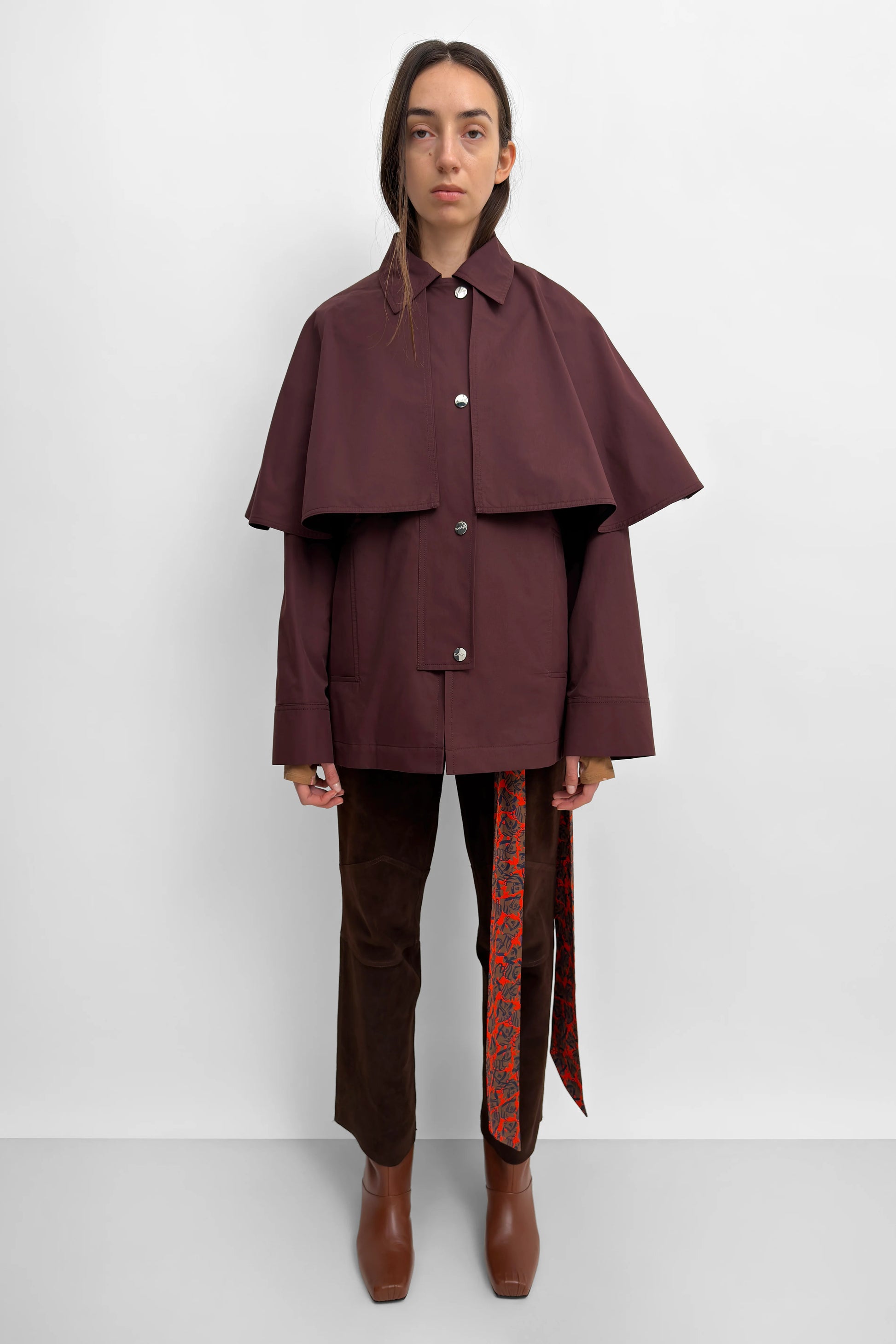 A model stands against a light gray studio background wearing a dark burgundy cape-style jacket with wide draped sleeves, dark brown trousers, and brown boots. A patterned red and green fabric hangs from the side of the pants.