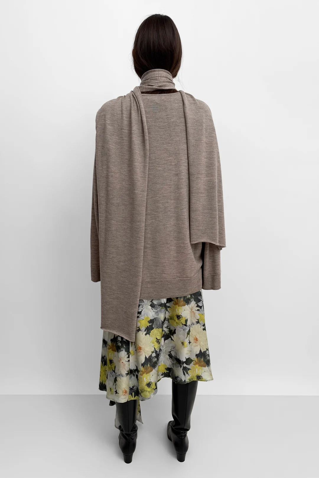 A model shown from the back wearing a taupe knit wrap or shawl draped over a floral print skirt, paired with black boots, against a plain white studio background.