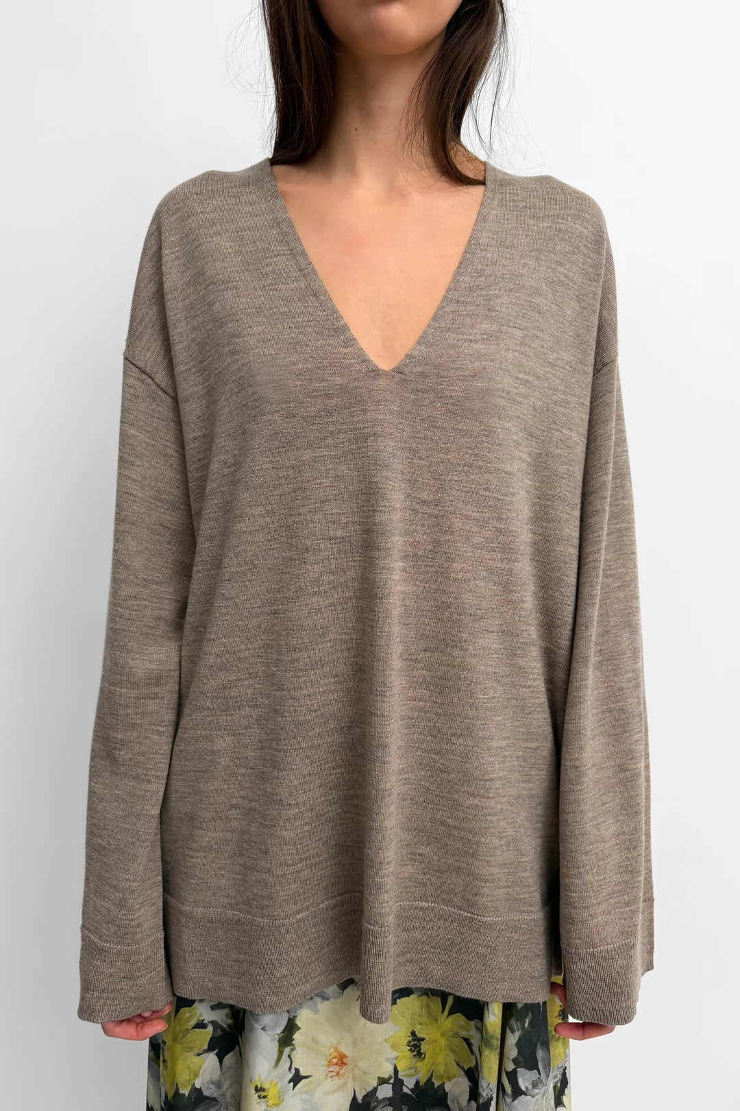 A woman wearing a taupe-brown long-sleeve knit sweater with a deep V-neck, paired with a floral skirt, against a plain light gray background.
