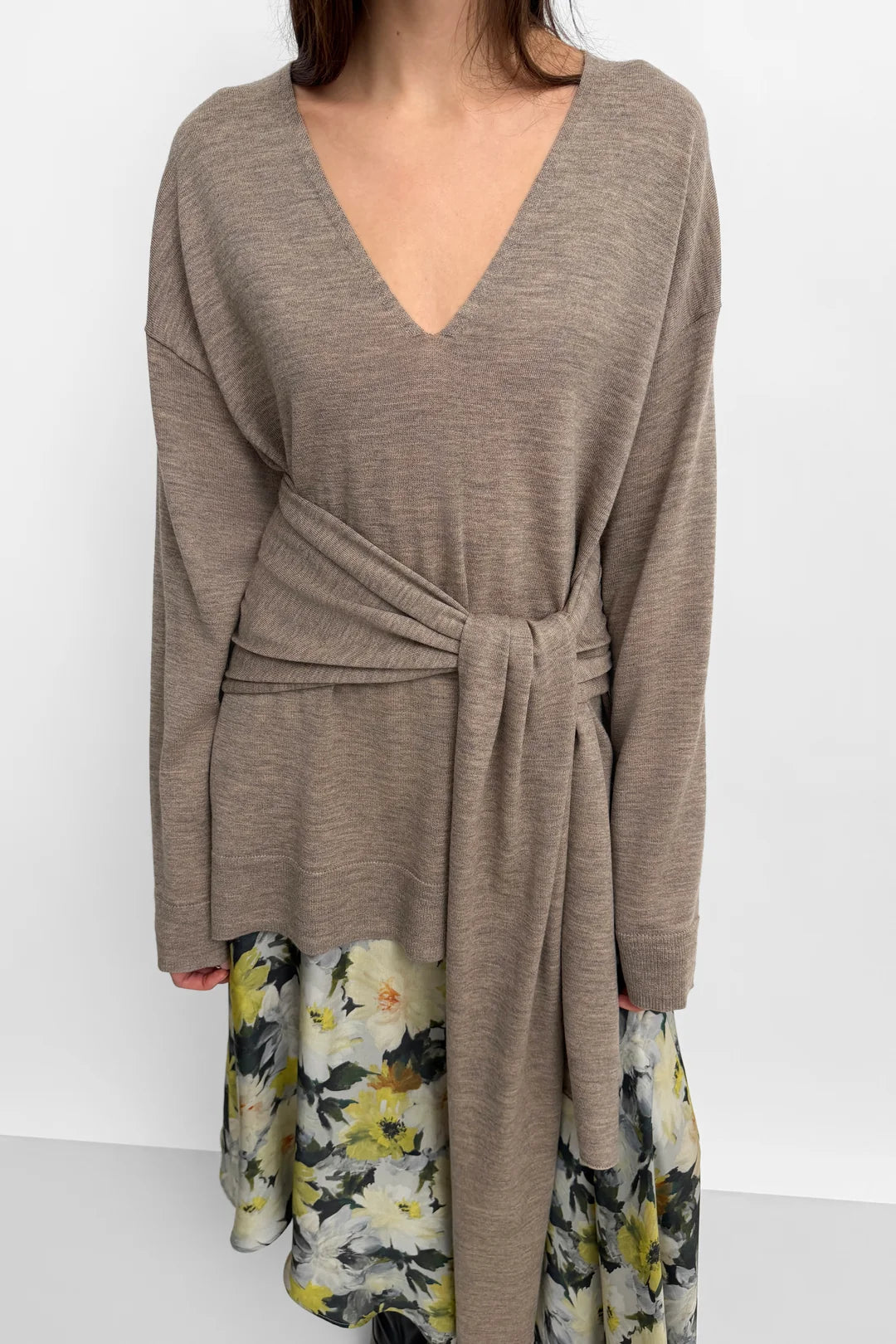 A woman wearing a taupe-brown long-sleeve wrap sweater with a V-neck and tie waist, layered over a floral print skirt, against a plain white background.