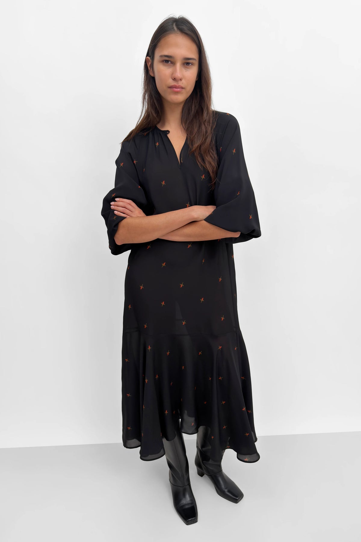 A woman modeling a black midi dress with a small orange-brown floral pattern, paired with black boots, against a plain white studio background.