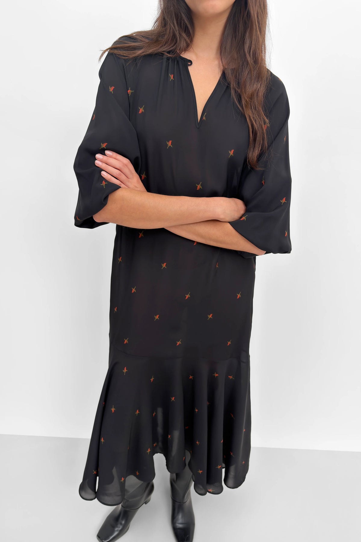 A woman wearing a black midi dress with small orange embroidered motifs, styled with black ankle boots against a plain white studio background.