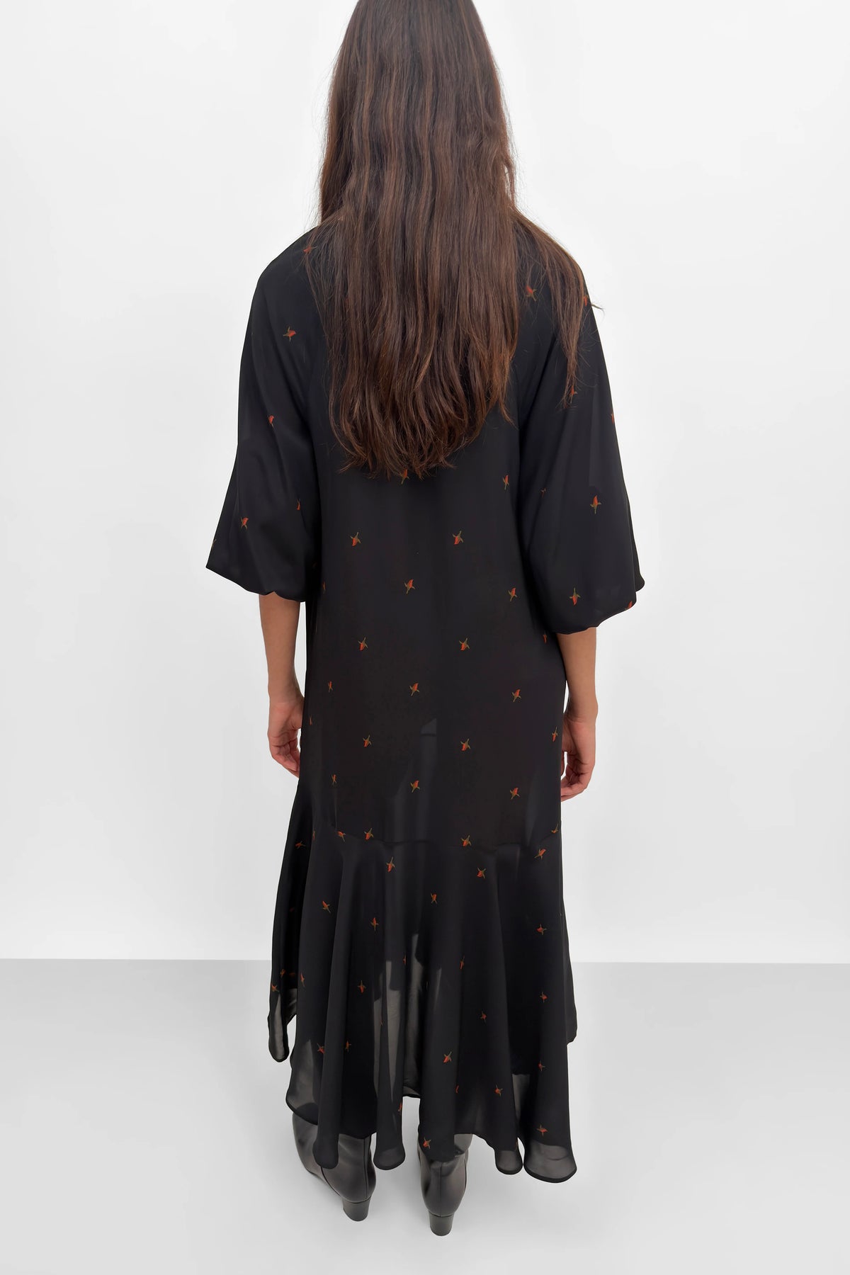 A woman viewed from behind wearing a black midi dress with three-quarter sleeves and a subtle orange pattern, standing against a plain white studio background.