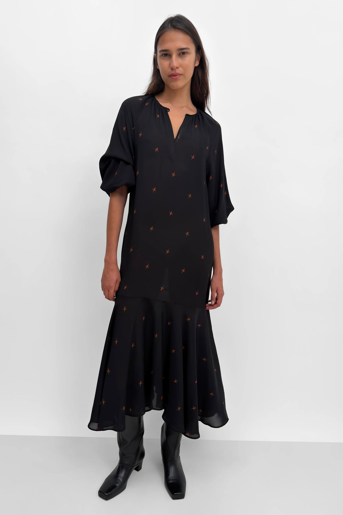 A woman modeling a black midi dress with small orange embroidered details, styled with black boots against a plain white studio background.