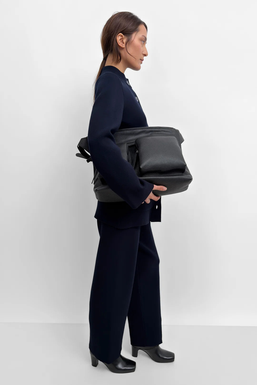 A woman in a dark navy blue suit holding a black leather messenger bag against a clean white studio background.