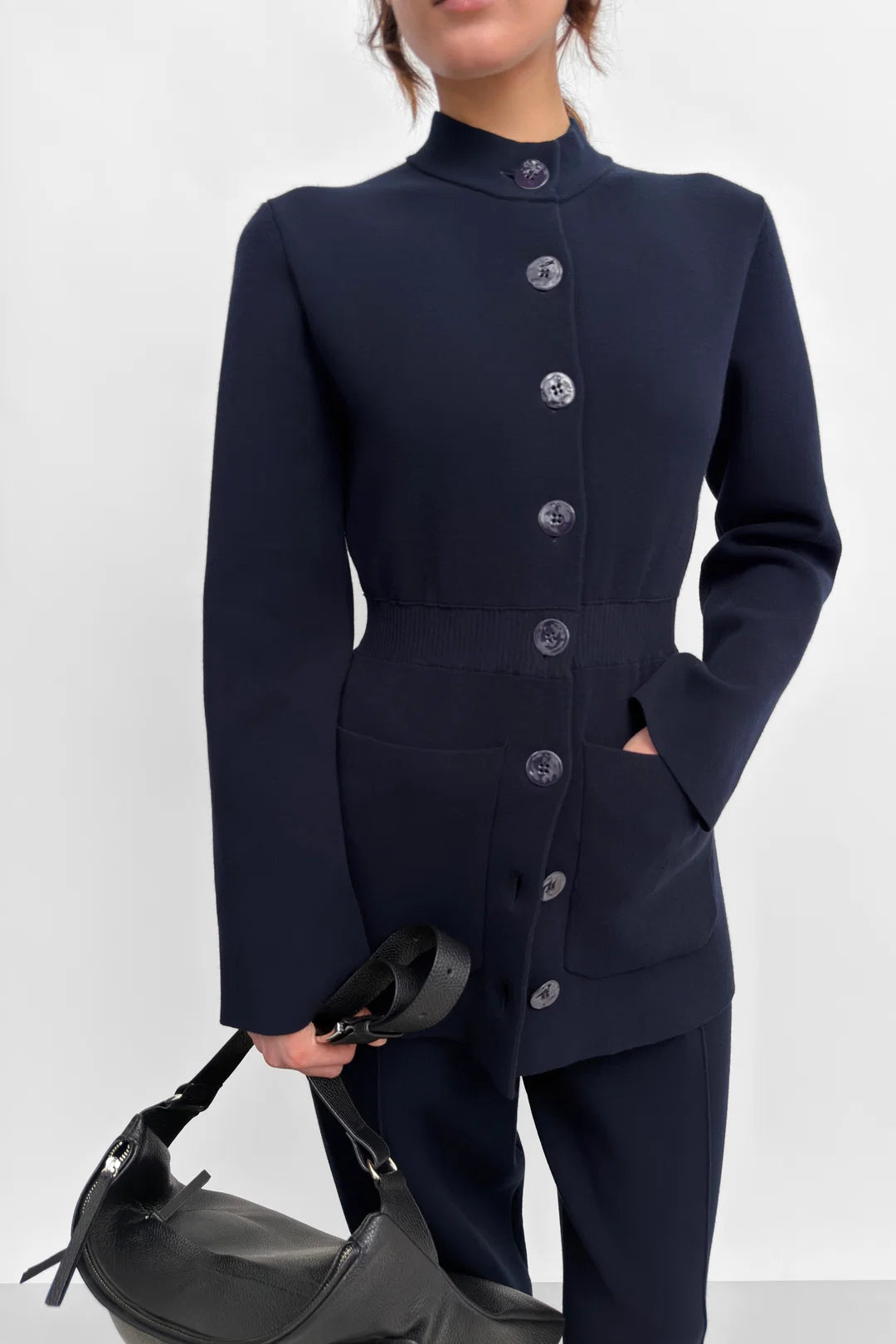 A model wearing a dark navy blue tailored jacket with matching trousers, featuring a high mock neck, structured shoulders, and a front button closure. The model is holding a black leather handbag against a plain light gray studio background.
