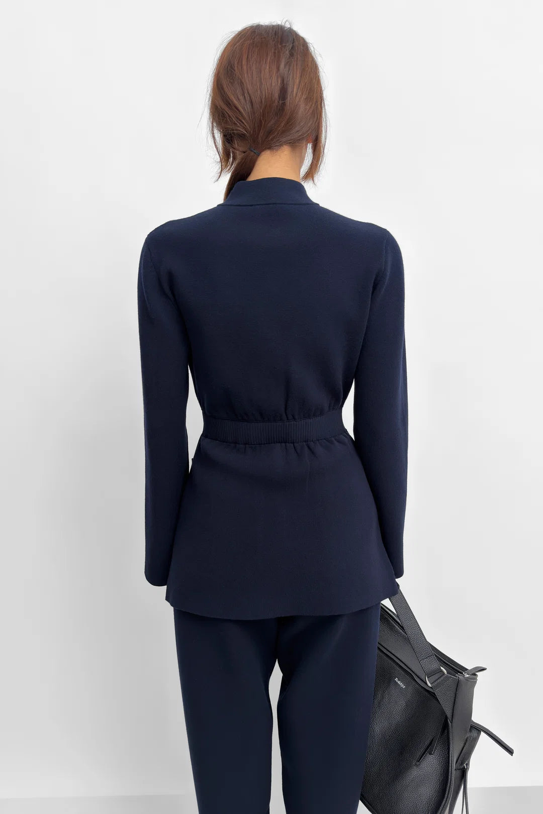 A woman shown from the back wearing a navy blue tailored blazer with a defined waist and matching navy trousers, standing against a plain light gray wall and holding a black handbag.