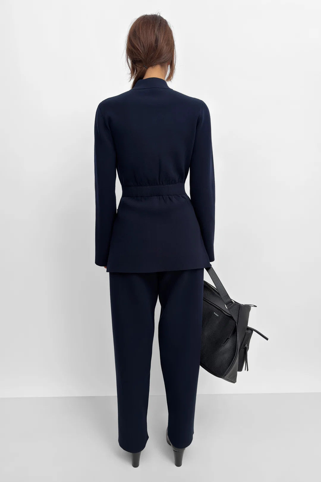 A model shown from the back wearing a dark navy blue tailored suit, featuring a belted blazer and matching trousers, holding a black leather handbag against a clean white studio background.