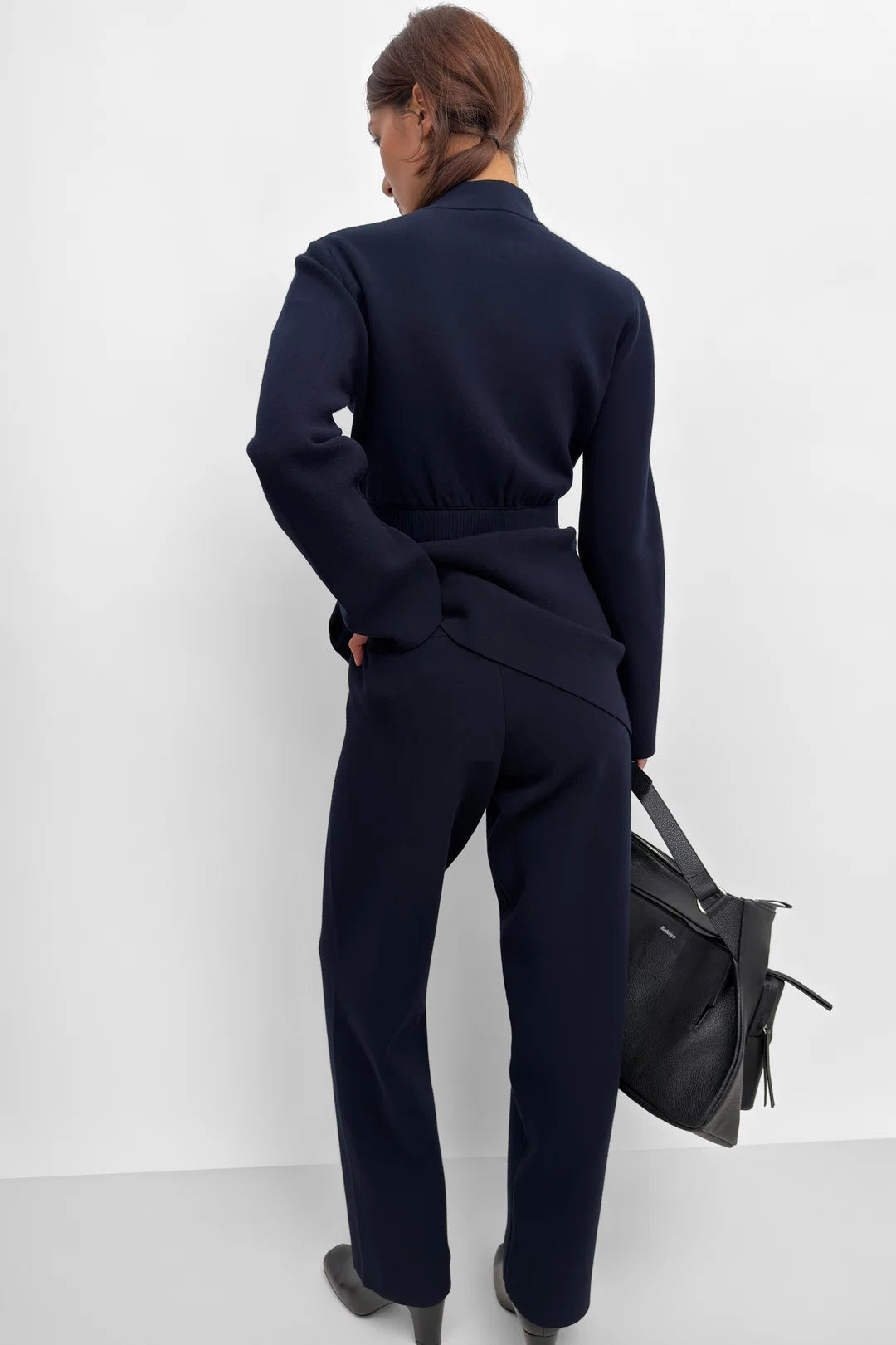 A woman shown from the back wearing a dark navy blue belted jacket and matching trousers, holding a black leather handbag against a plain white studio background.