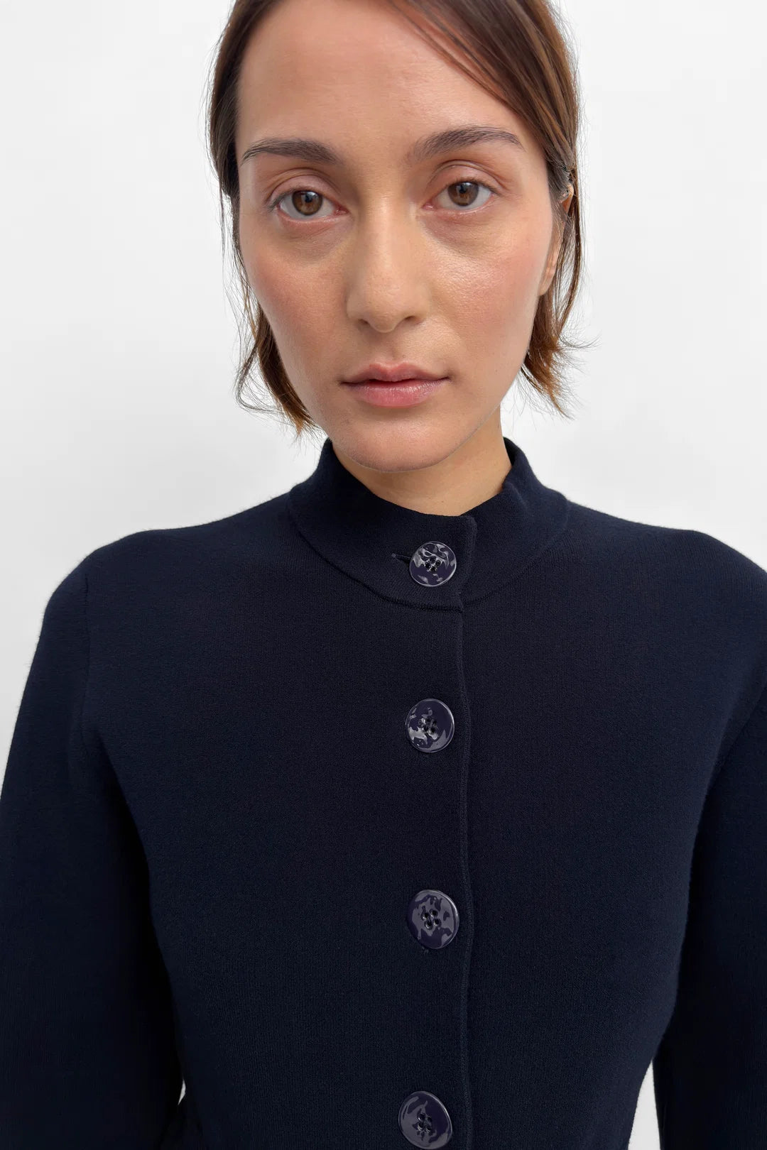 A close-up shot of a model wearing a dark navy blue knit top with a mock neck and decorative dark buttons down the front, set against a plain light gray background.