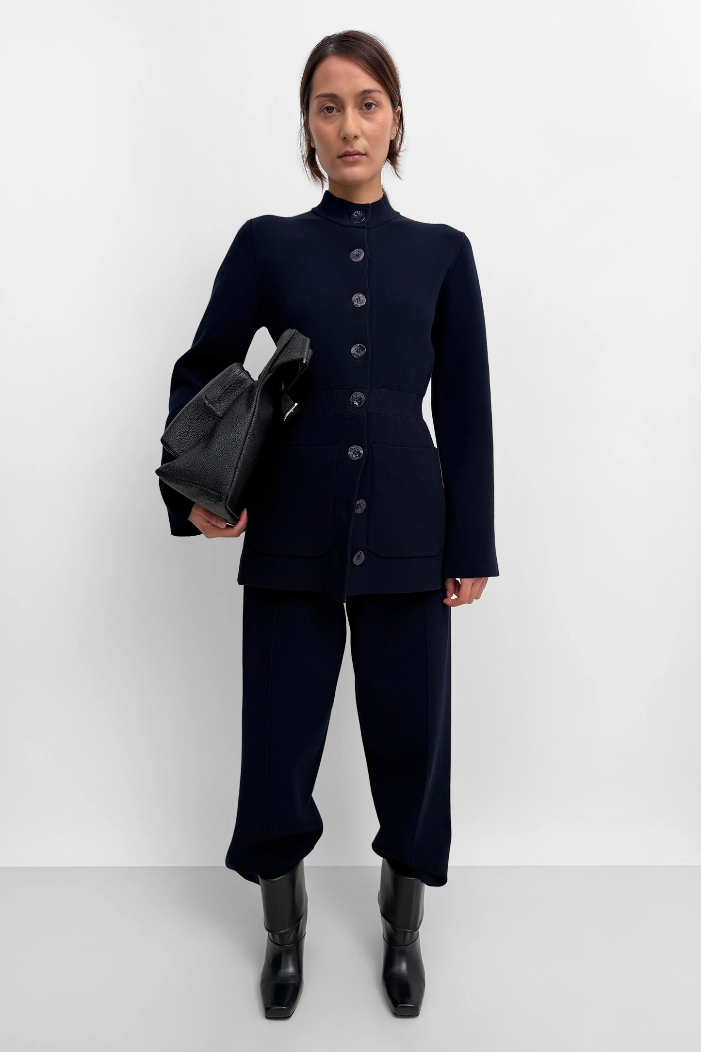 A model stands against a light gray studio background wearing a dark navy blue tailored set consisting of a long-sleeve button-front jacket with a high mock neck and matching wide-leg trousers. She is holding a black handbag and wearing black pointed boots.