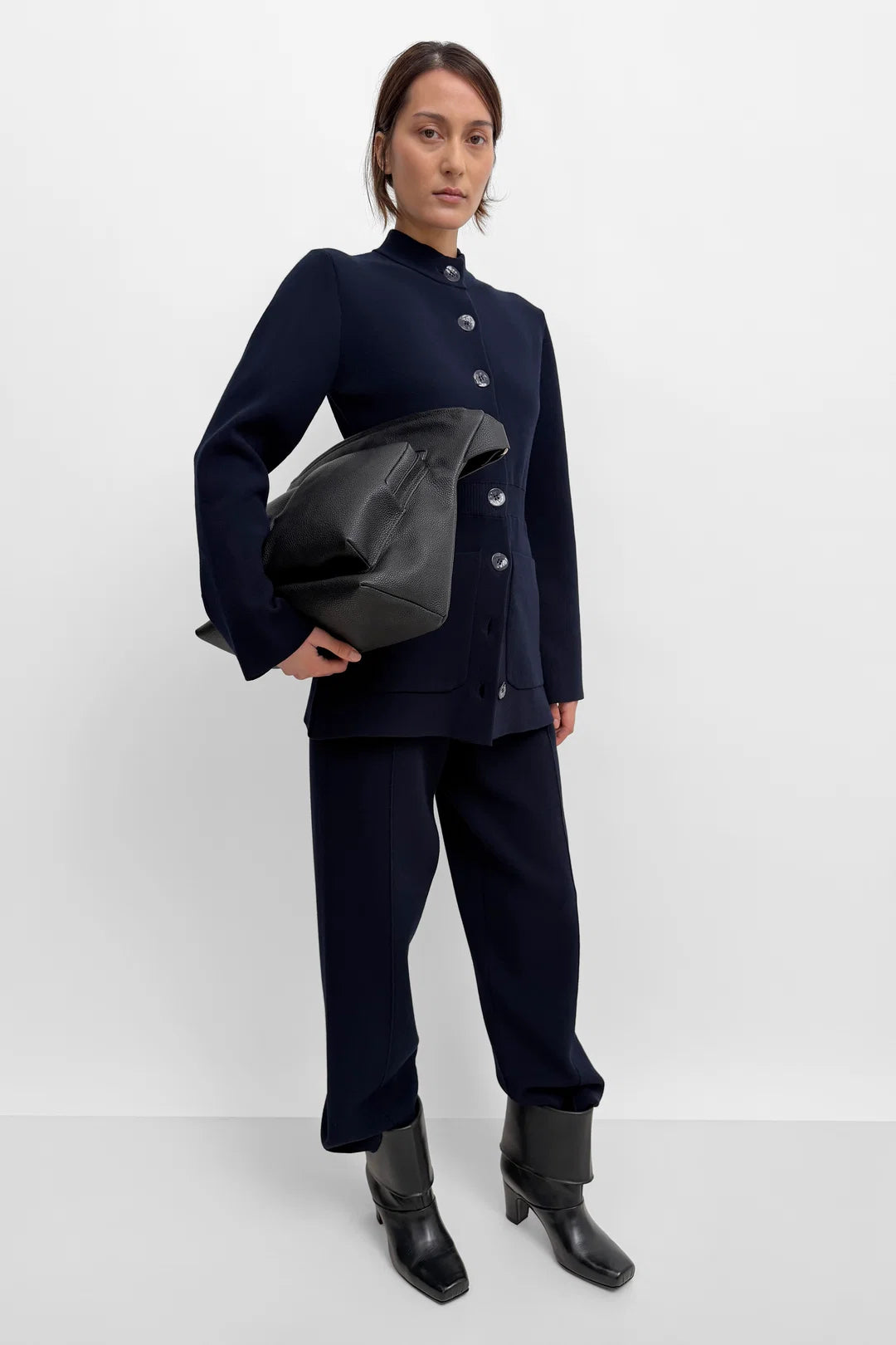 A model stands against a light gray studio background wearing a dark navy blue two-piece outfit consisting of a long-sleeve button-front jacket and matching wide-leg trousers, paired with black ankle boots and holding a black leather bag.