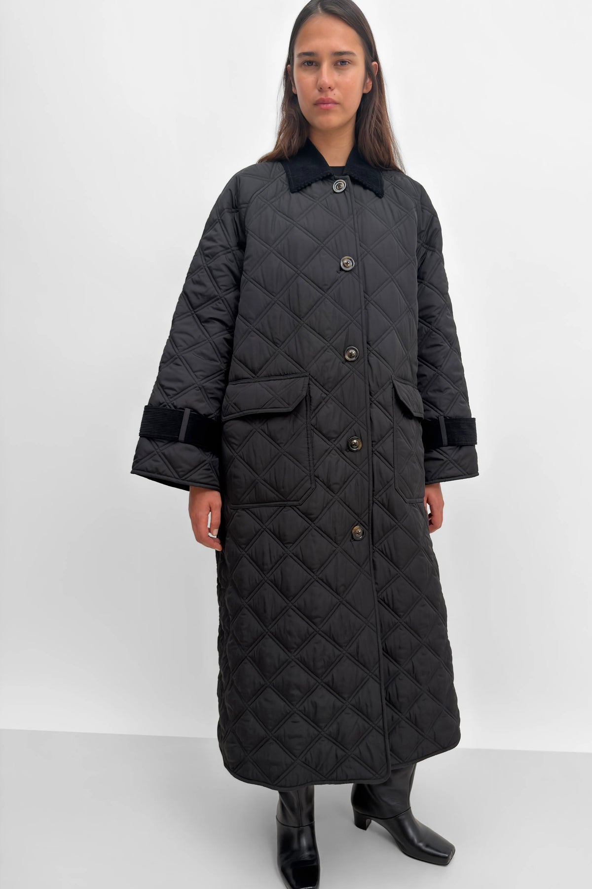 A model wearing a long black diamond-quilted coat with black velvet trim on the collar, cuffs, and pockets, paired with black leather boots, against a plain light gray studio background.