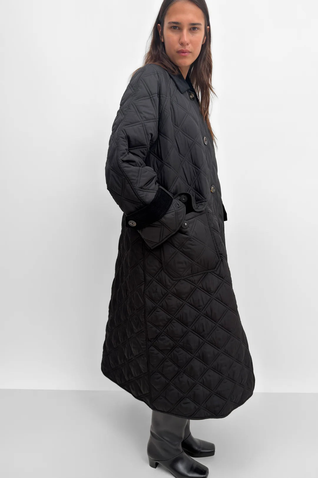 A model wearing a long black diamond-quilted coat with black leather-like trim on the cuffs, paired with black boots, against a plain white studio background.