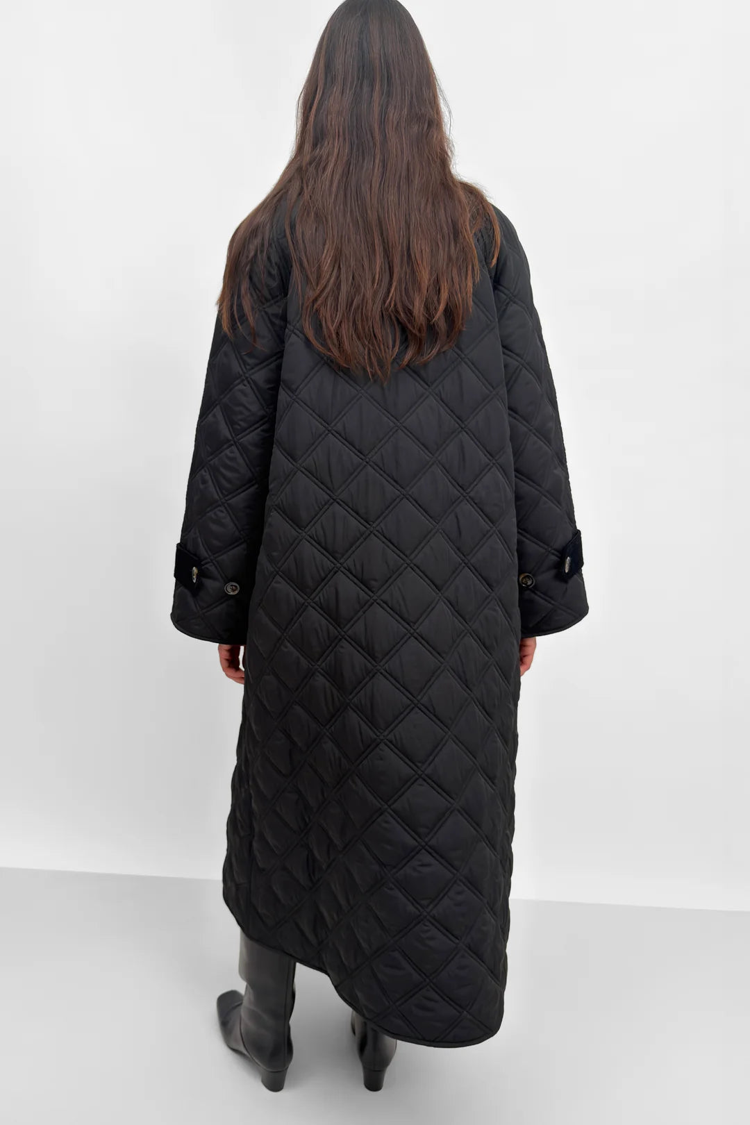 A woman viewed from behind wearing a long black quilted coat with diamond stitching, shown against a plain light gray studio background.
