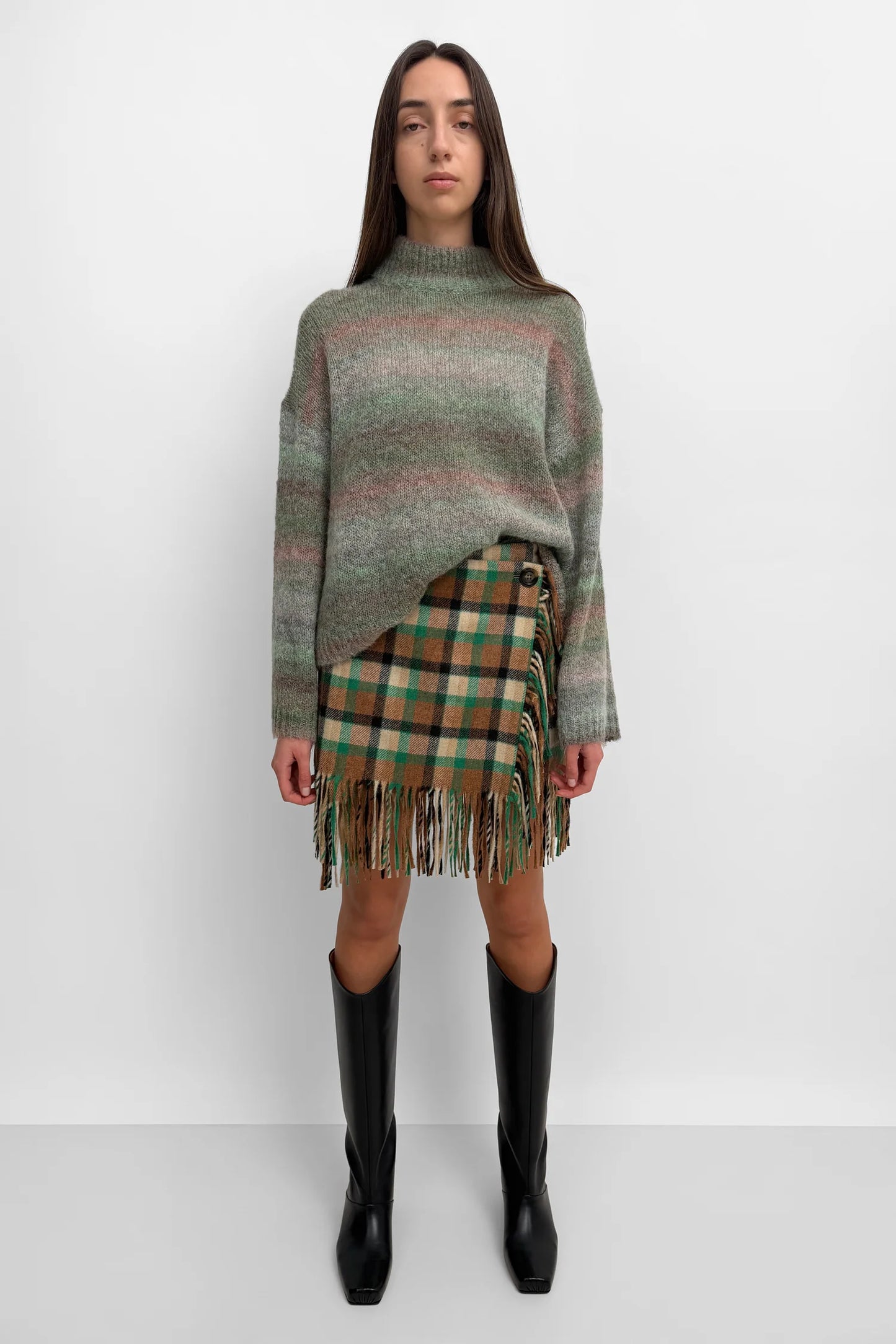 A model stands against a light gray studio background wearing a fuzzy striped knit sweater in muted green, beige, and pink tones, paired with a green and tan plaid fringe mini skirt and black knee-high boots.