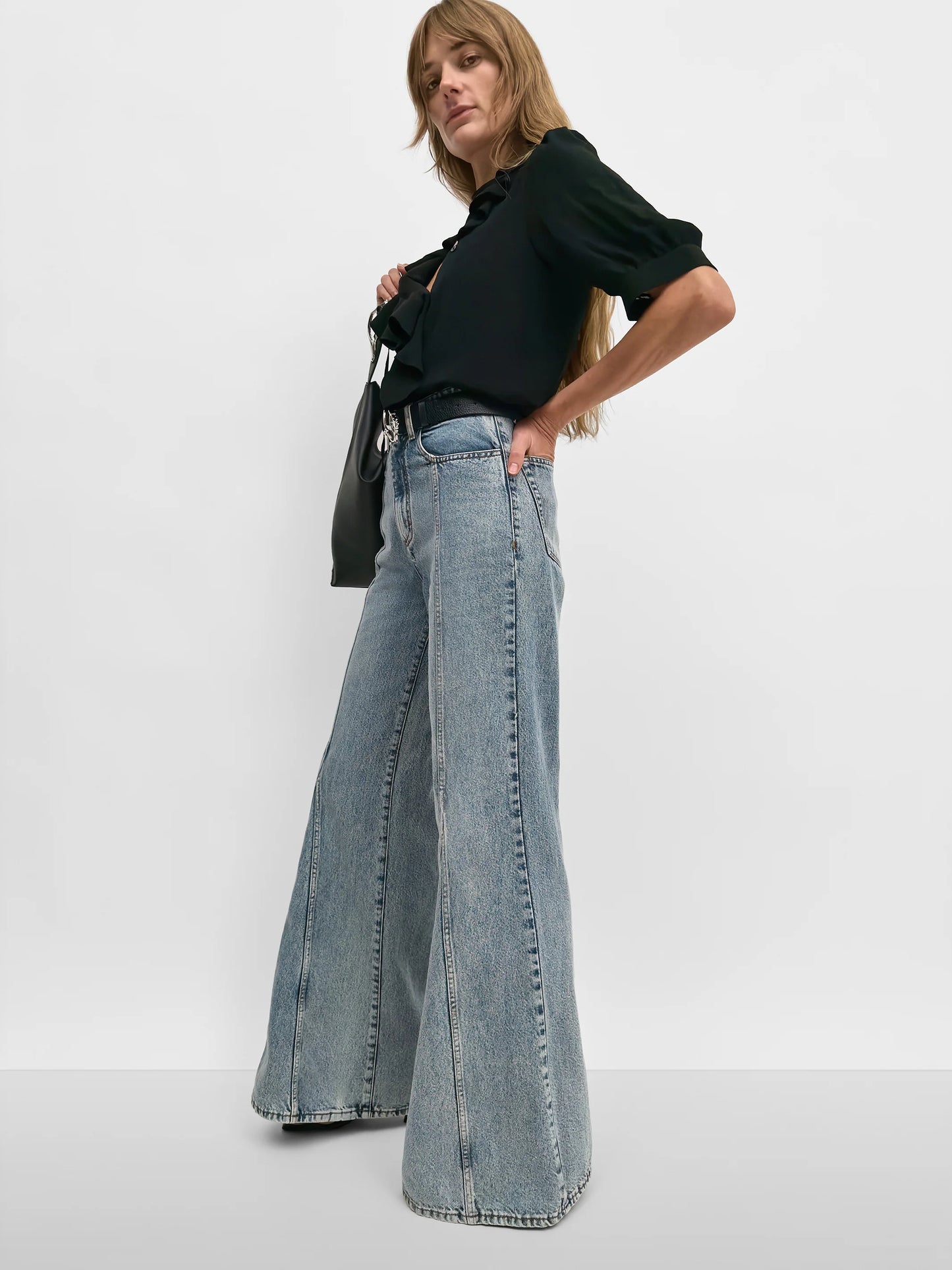 A woman modeling light-wash wide-leg denim jeans with a black ruffled blouse, standing against a plain light gray studio background.