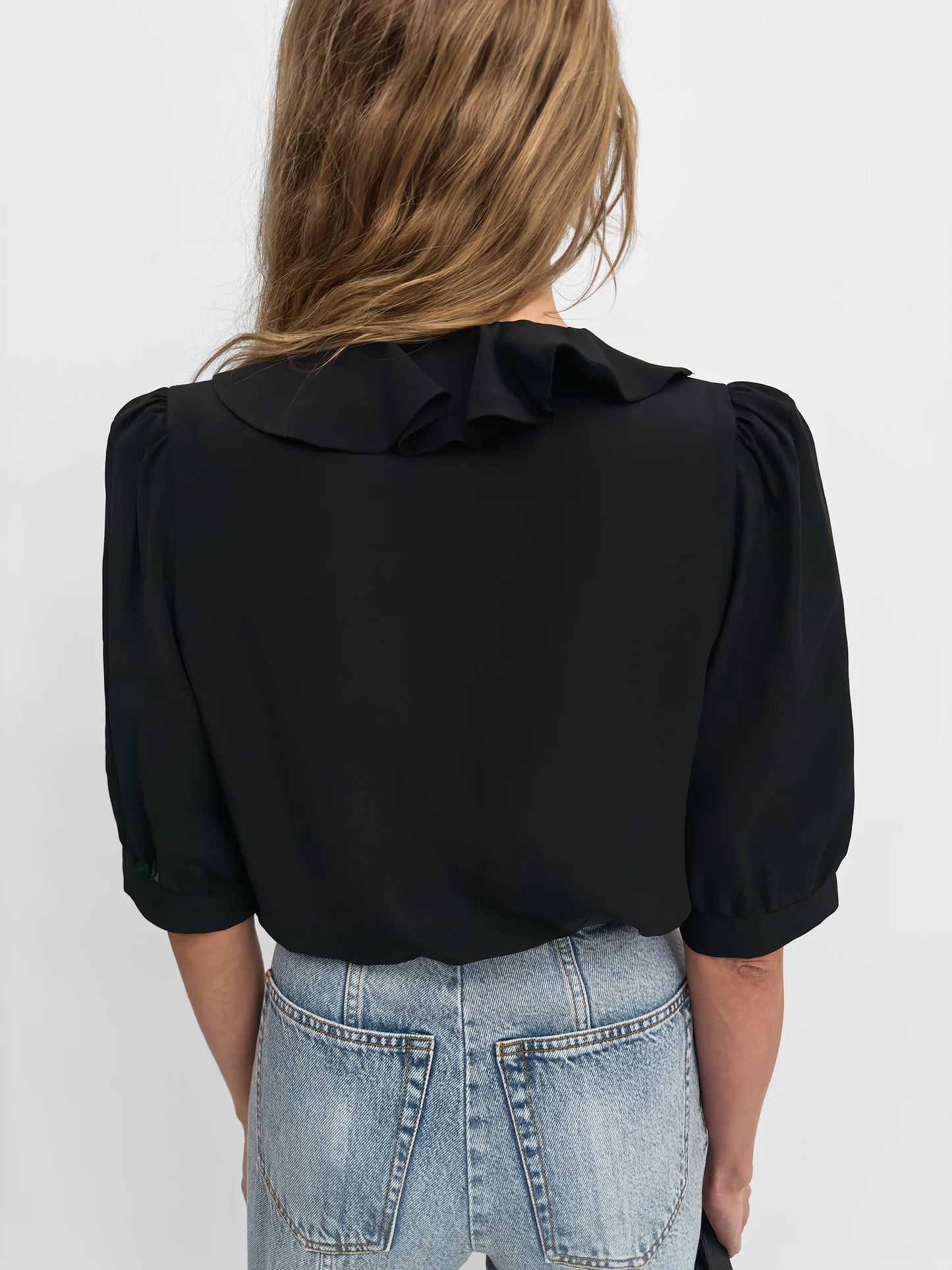 A woman viewed from the back wearing a black blouse with a ruffled collar and puff sleeves, paired with light blue jeans, against a plain light gray background.