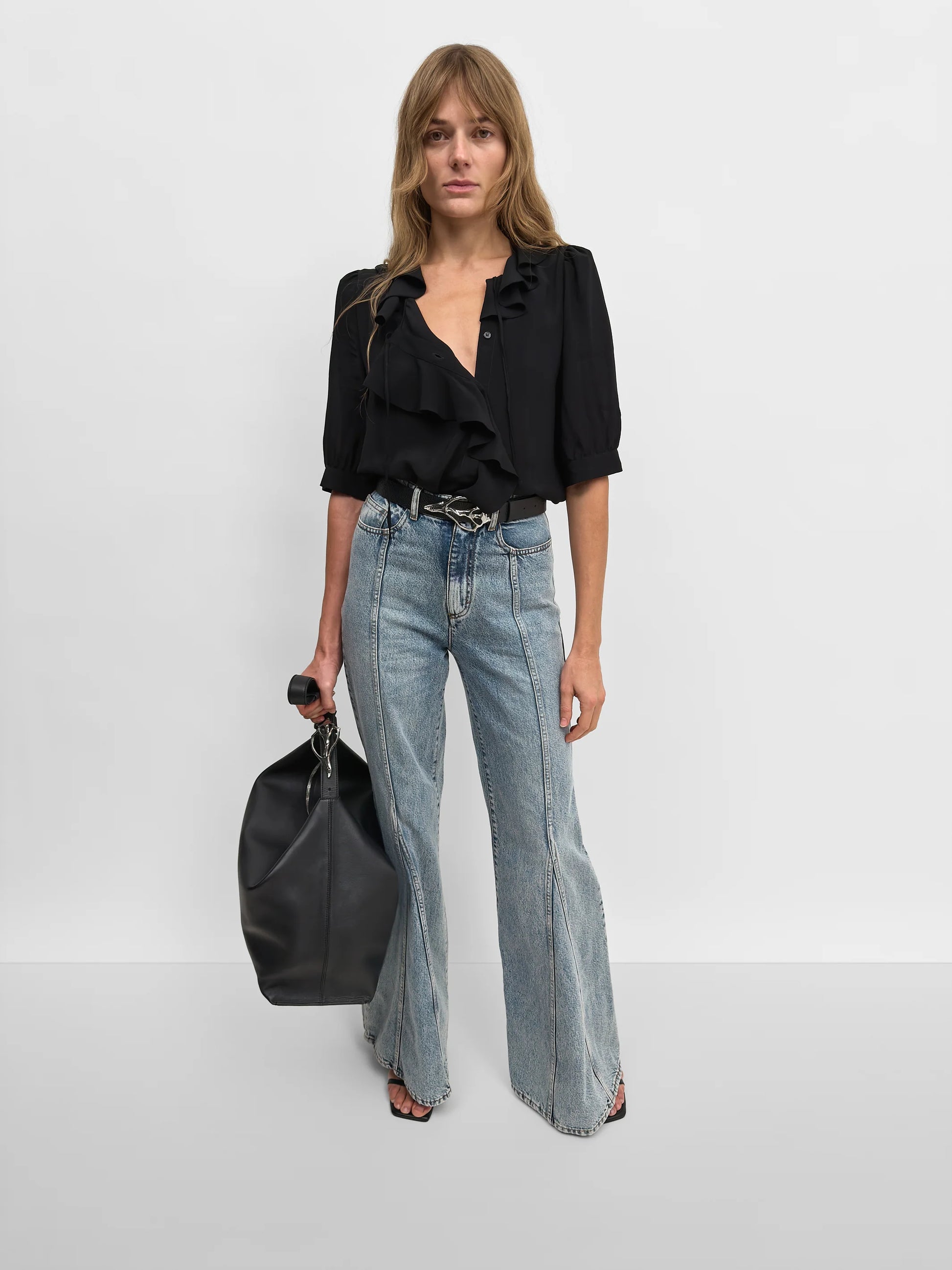 A woman modeling a black ruffled blouse, light-wash wide-leg denim jeans, and a black leather duffel bag against a plain light gray studio background.