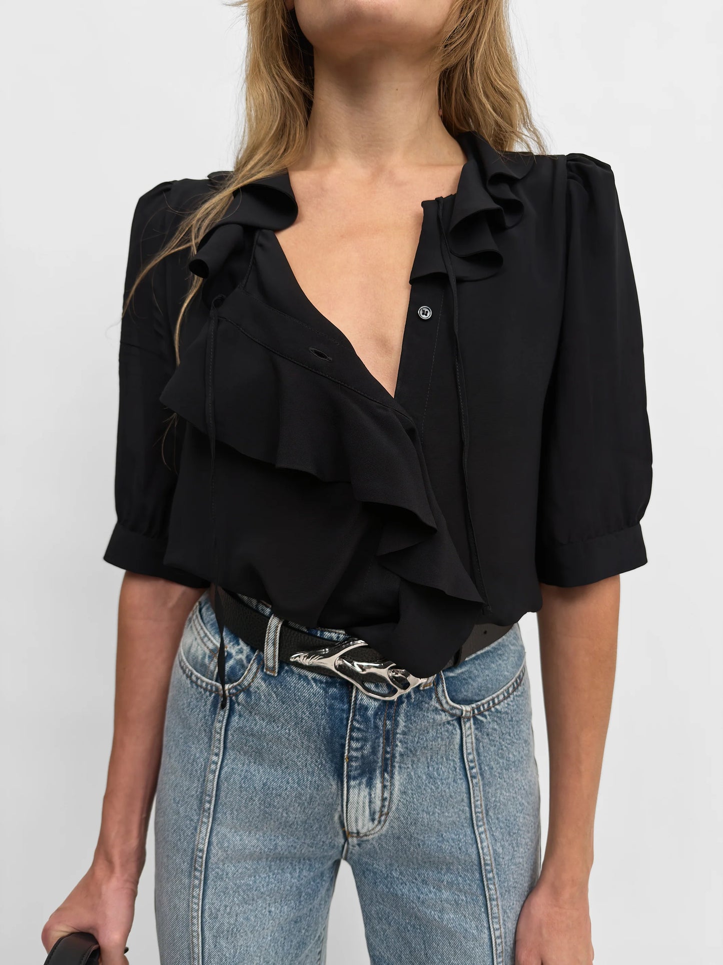A woman wearing a black ruffled blouse with puff sleeves and a deep V-neckline, paired with light-wash blue jeans and a black belt.