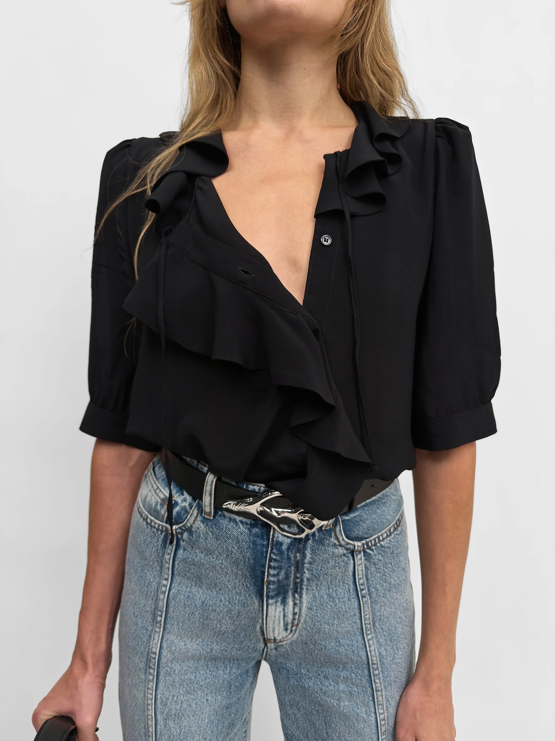 A woman wearing a black ruffled blouse with puff sleeves and a deep V-neckline, paired with light-wash blue jeans and a black belt.