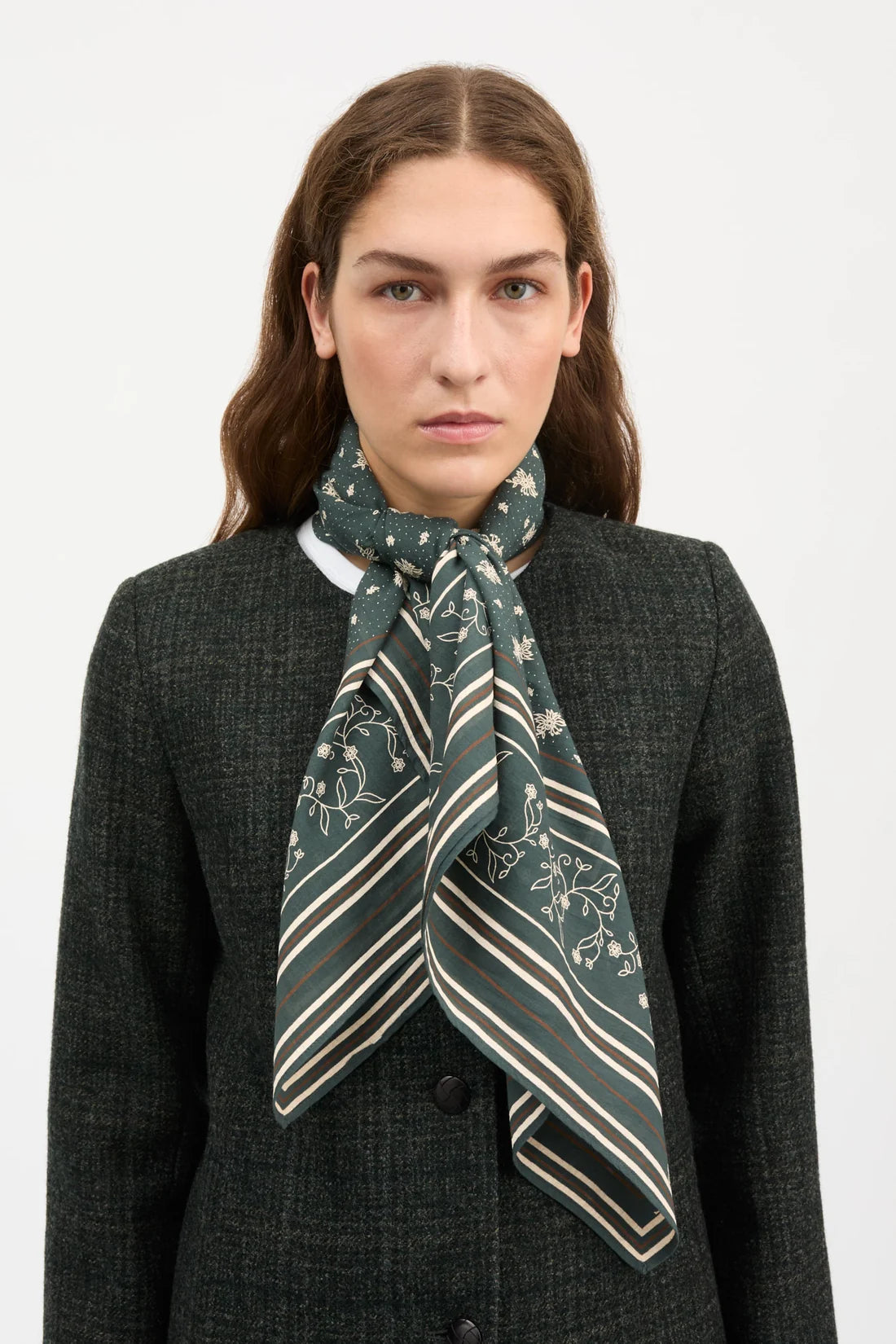 A woman wearing a dark green silk scarf with white floral and striped patterns, styled around her neck over a dark gray blazer, against a plain light gray background.