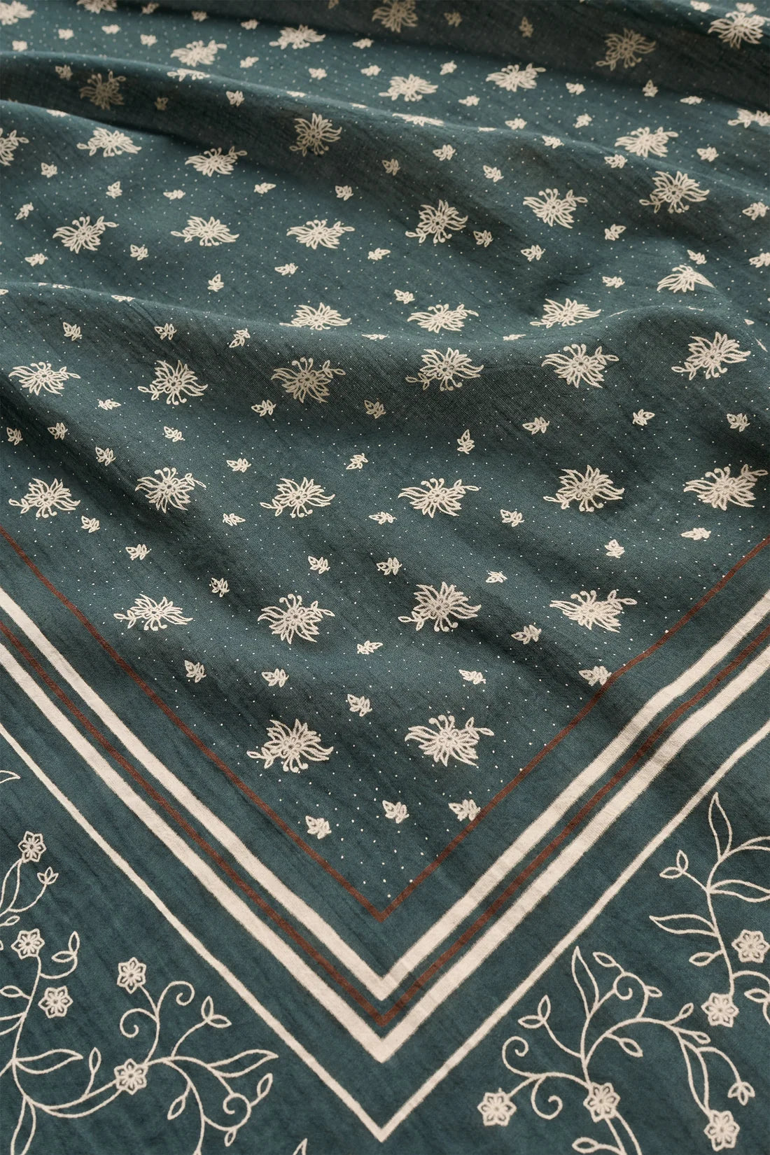 A close-up of a dark teal-green fabric with a repeating cream floral block print and a decorative cream border with thin red accent lines.
