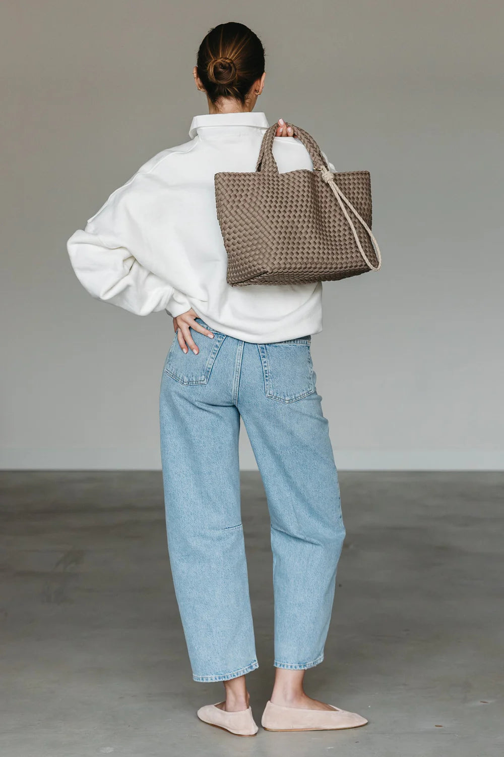 Person wearing a white sweater and light blue jeans, holding a woven brown bag.