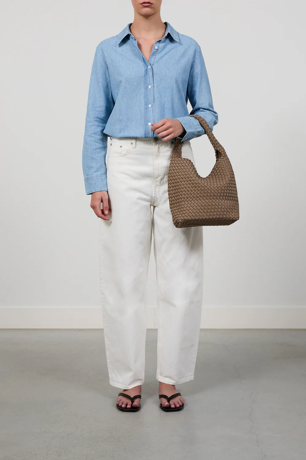 A model wearing a light blue long-sleeve button-up shirt, white wide-leg jeans, and black sandals, holding a brown woven leather shoulder bag against a neutral studio background.