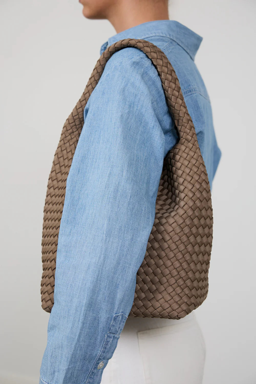 A brown woven leather shoulder bag with a wide strap, worn over the shoulder of a person wearing a light blue denim shirt, shown from a side profile view against a plain light gray background.