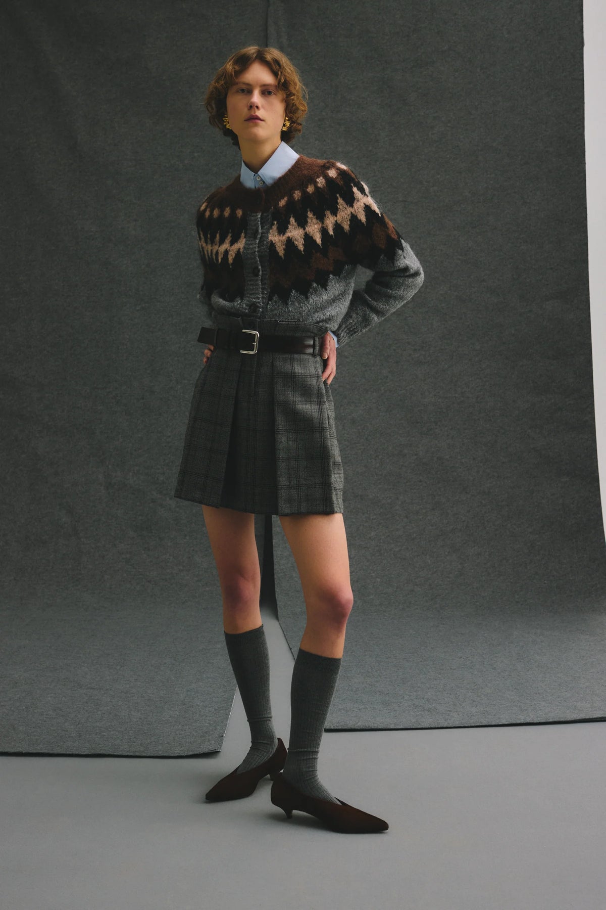 A model stands against a dark gray studio backdrop wearing a gray knit sweater with a brown and beige geometric pattern, a gray plaid pleated mini skirt, a light blue collared shirt underneath, dark gray knee-high socks, and dark brown pointed-toe loafers.