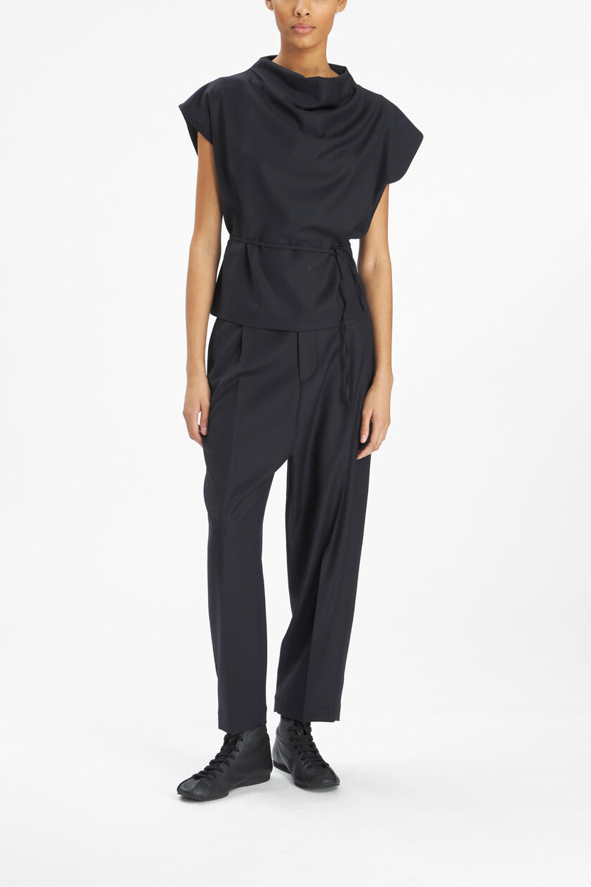 A model wearing a black draped cowl-neck jumpsuit with short cap sleeves, a cinched waist, and wide-leg trousers, paired with black lace-up shoes, against a plain white studio background.
